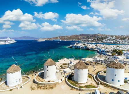 A photo of the well-known windmills in Mykonos, Greece with a view of the different shades of blue in the sea and mountains in the background.